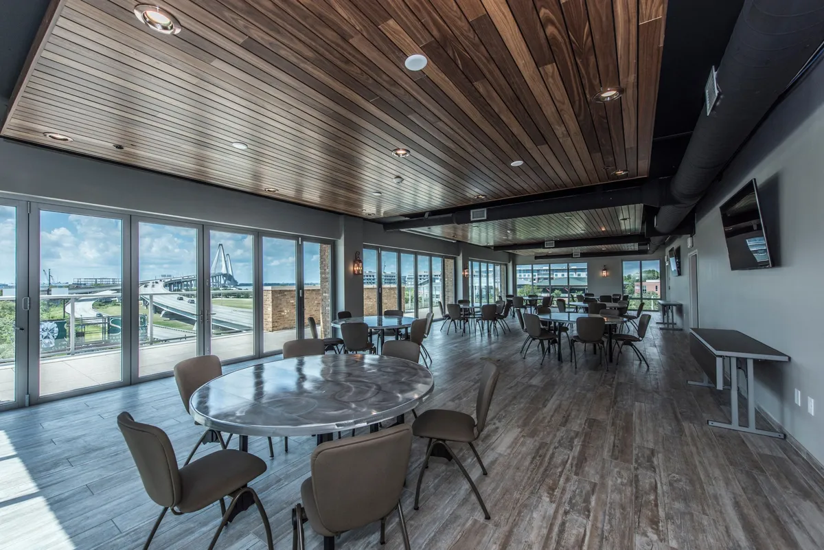 A bright, contemporary interior gathering space with large windows flooding the room with natural light. The area features round tables perfect for informal meetings, networking, or social gatherings, and offers views of the outdoors, blending the themes of productivity and relaxation.