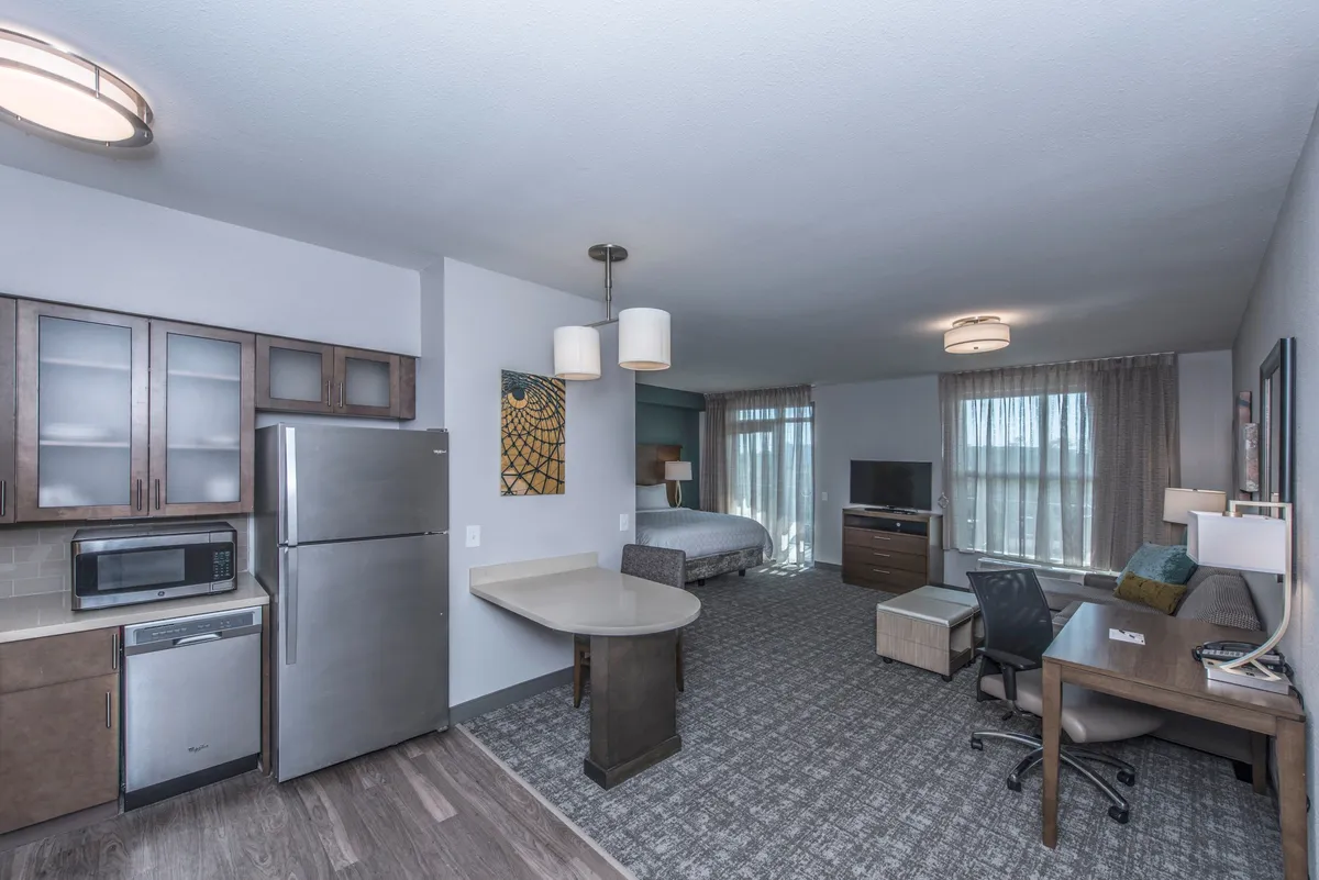 A well-lit studio interior featuring a king bed with plush linens, a compact kitchen area, a dedicated workspace with a desk and chair, and a flat-screen TV. Soft lighting and tasteful decor make the space feel inviting and multifunctional.