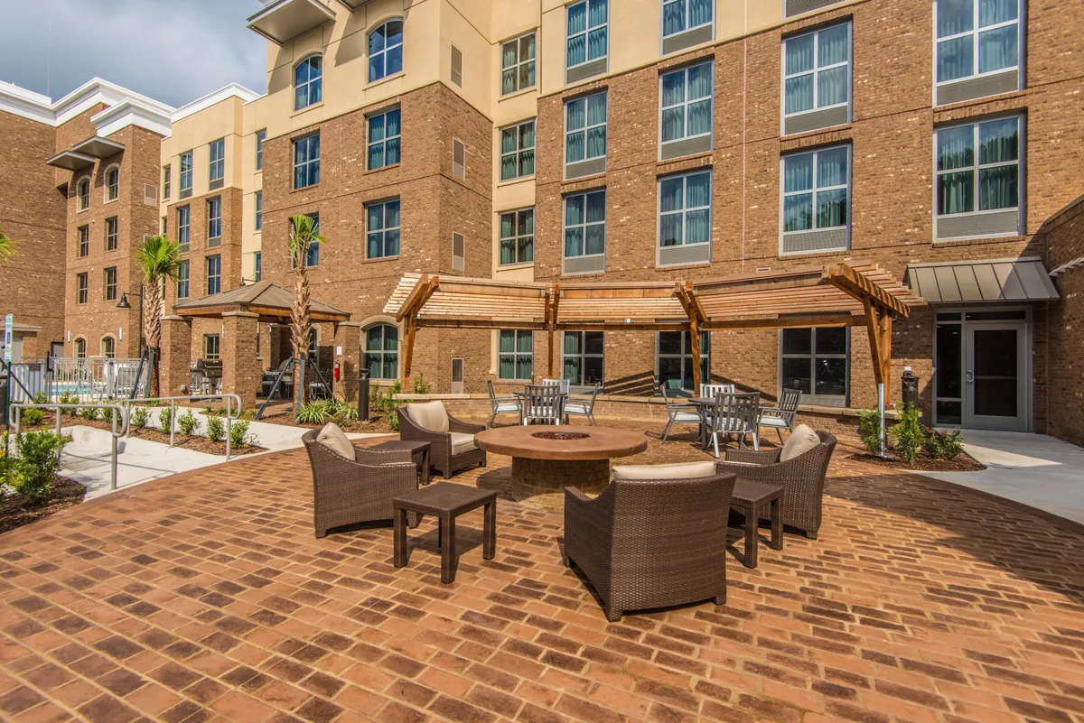An exterior communal area centered around a fire pit, surrounded by comfortable seating and landscaped gardens. The scene suggests a relaxed environment ideal for after-work socializing or informal business networking under the open sky.
