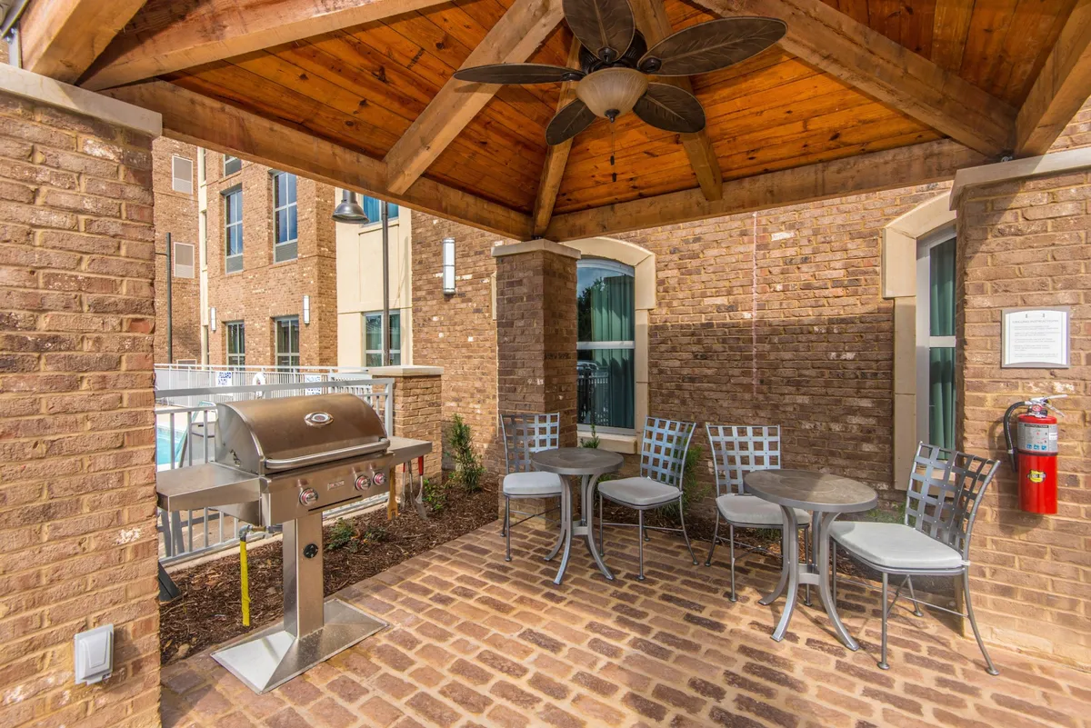 An inviting semi-outdoor space with a barbecue grill, comfortable seating, a wooden ceiling, and exposed brick walls. The area is designed for communal dining, evening gatherings, and relaxed socializing, creating a bridge between indoor comfort and outdoor ambiance.