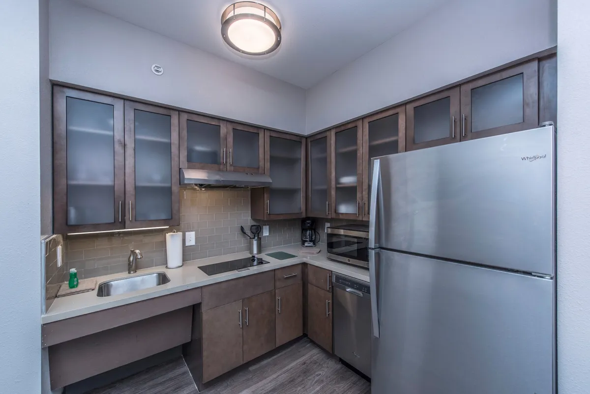 A contemporary studio kitchenette with a stainless steel refrigerator, sleek kitchen cabinets, and a generous sink set against a clean, modern backdrop. The space suggests easy meal prep and social cooking, and hints at the presence of appliances like a stove and microwave within the cabinetry.