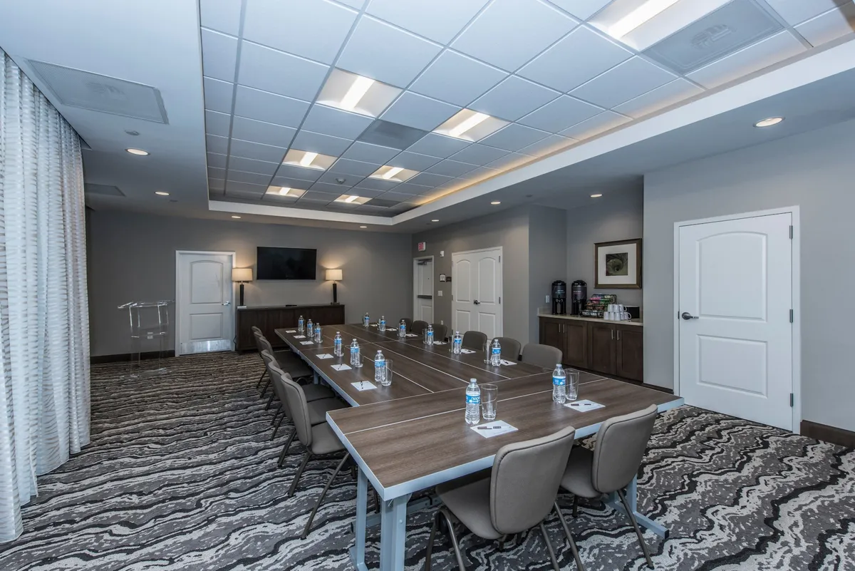 A sleek conference room features a long wooden table set with water bottles and a large television screen, ideal for business meetings, presentations, and collaborative work. The setup is professional yet inviting, suitable for productive sessions away from the office.