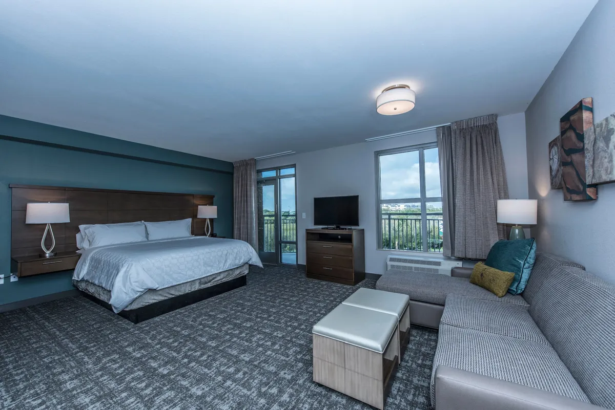 A serene studio scene with a comfortable king bed, a small sofa for lounging, a large window letting in natural light, and a television positioned for easy viewing. The decor is soft and relaxing, creating a peaceful retreat vibe.