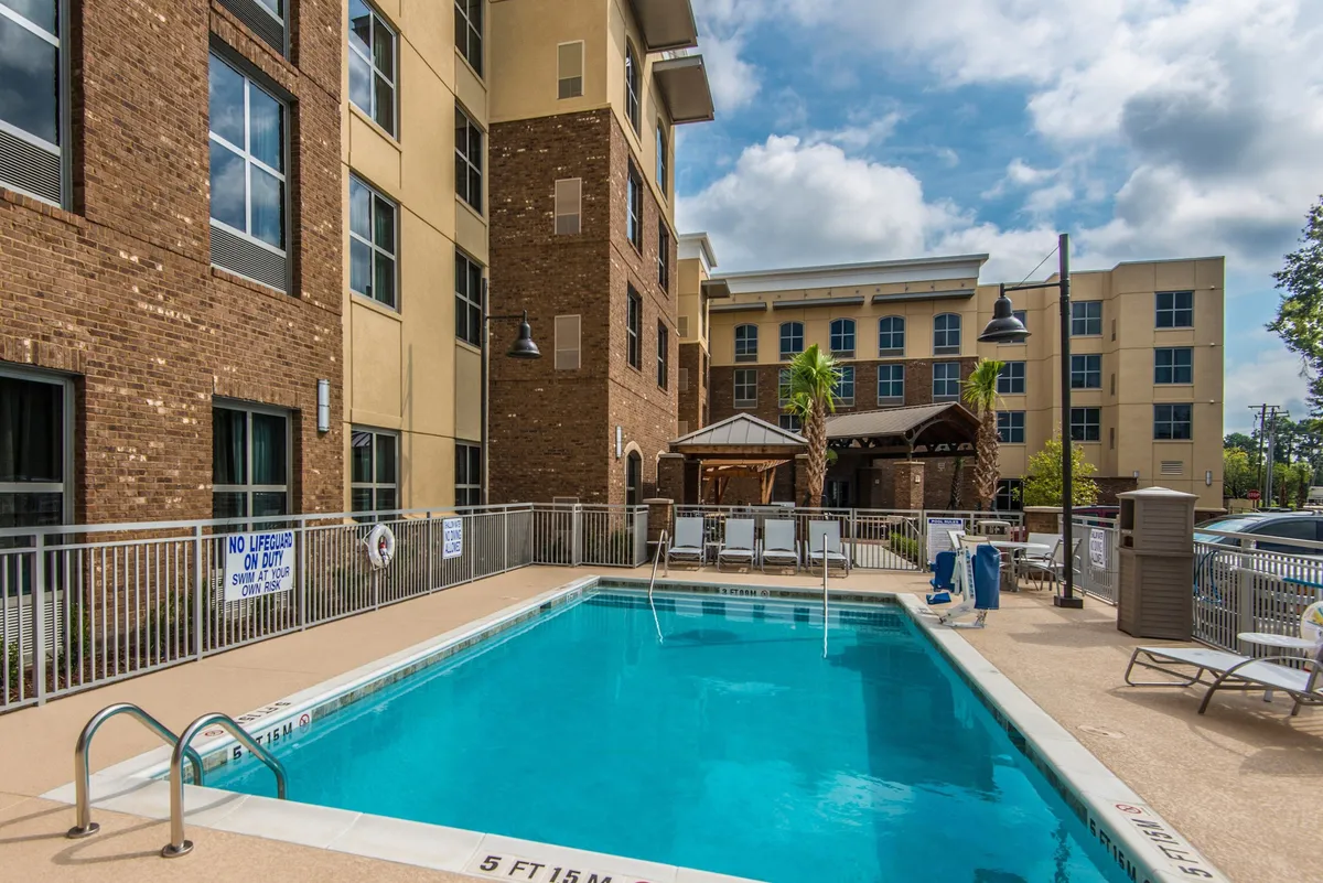 This exterior image features a sparkling swimming pool surrounded by lounge chairs, a cabana structure, and nearby buildings. The scene captures the pool area during golden hour, with a relaxed atmosphere and spaces for sunbathing and socializing.
