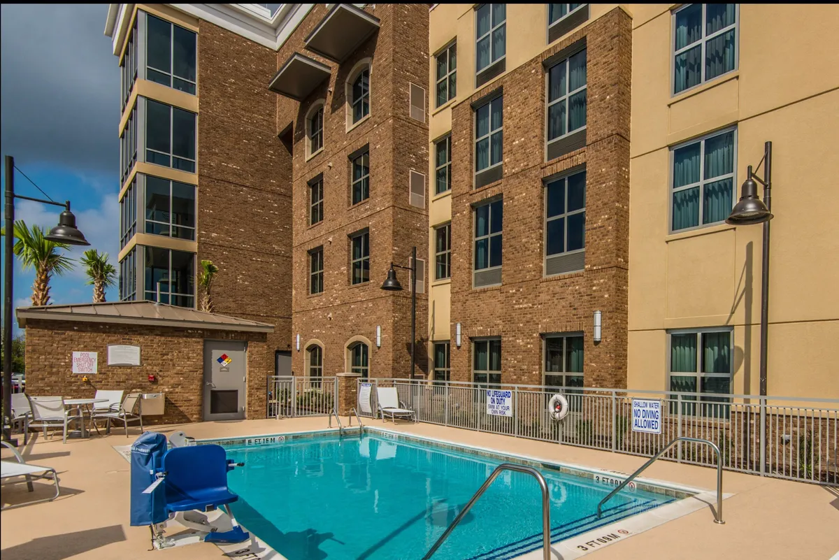 This exterior image features a sparkling swimming pool surrounded by a brick building and an array of inviting lounge chairs. The relaxed atmosphere is accentuated by the gentle light, hinting at sunset swims and social gatherings. The scene suggests a vibrant, welcoming space for unwinding after a day of exploration in Charleston.