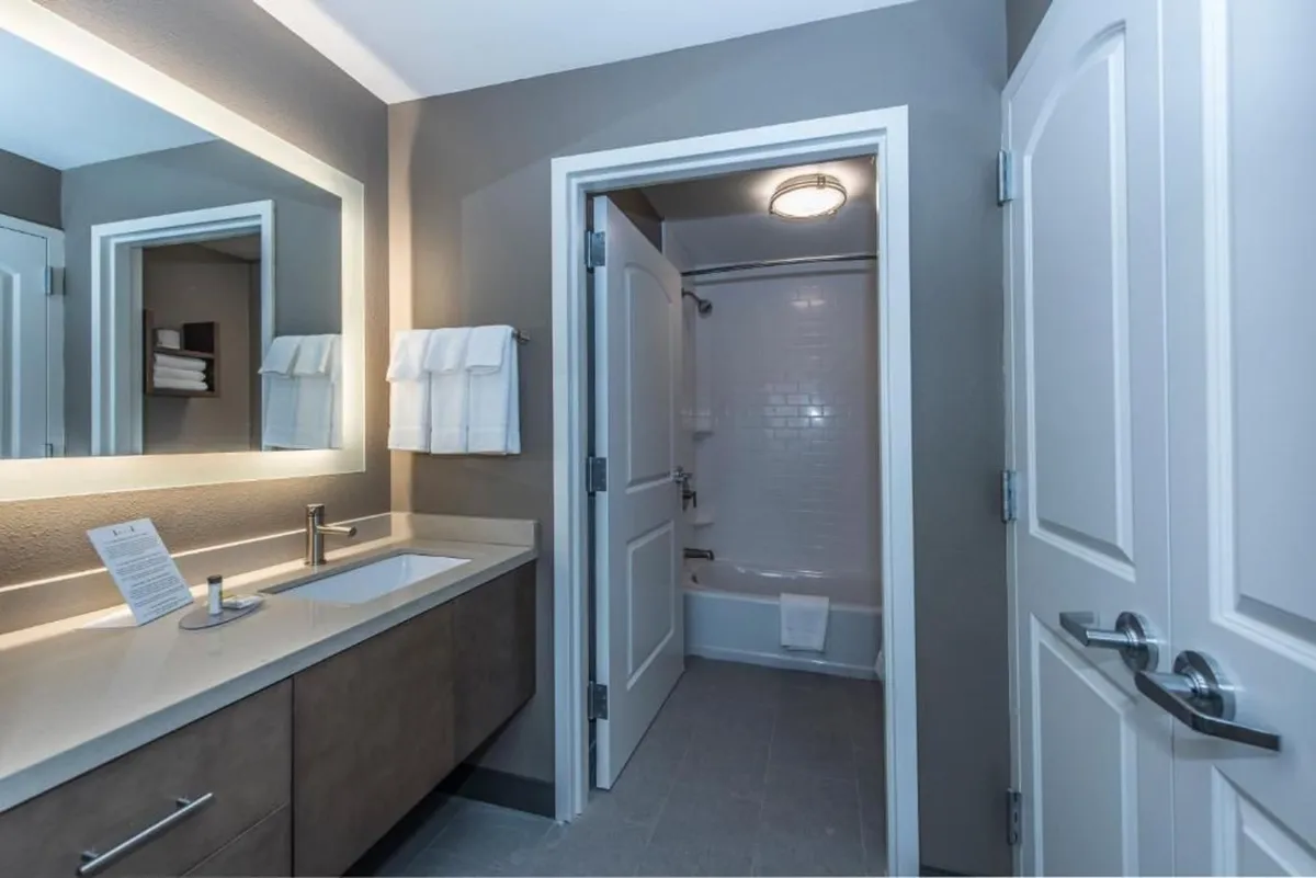The image showcases a modern, spacious bathroom with a large mirror, elegant sink area, and inviting walk-in shower—bathed in soft, serene light and designed for relaxation.