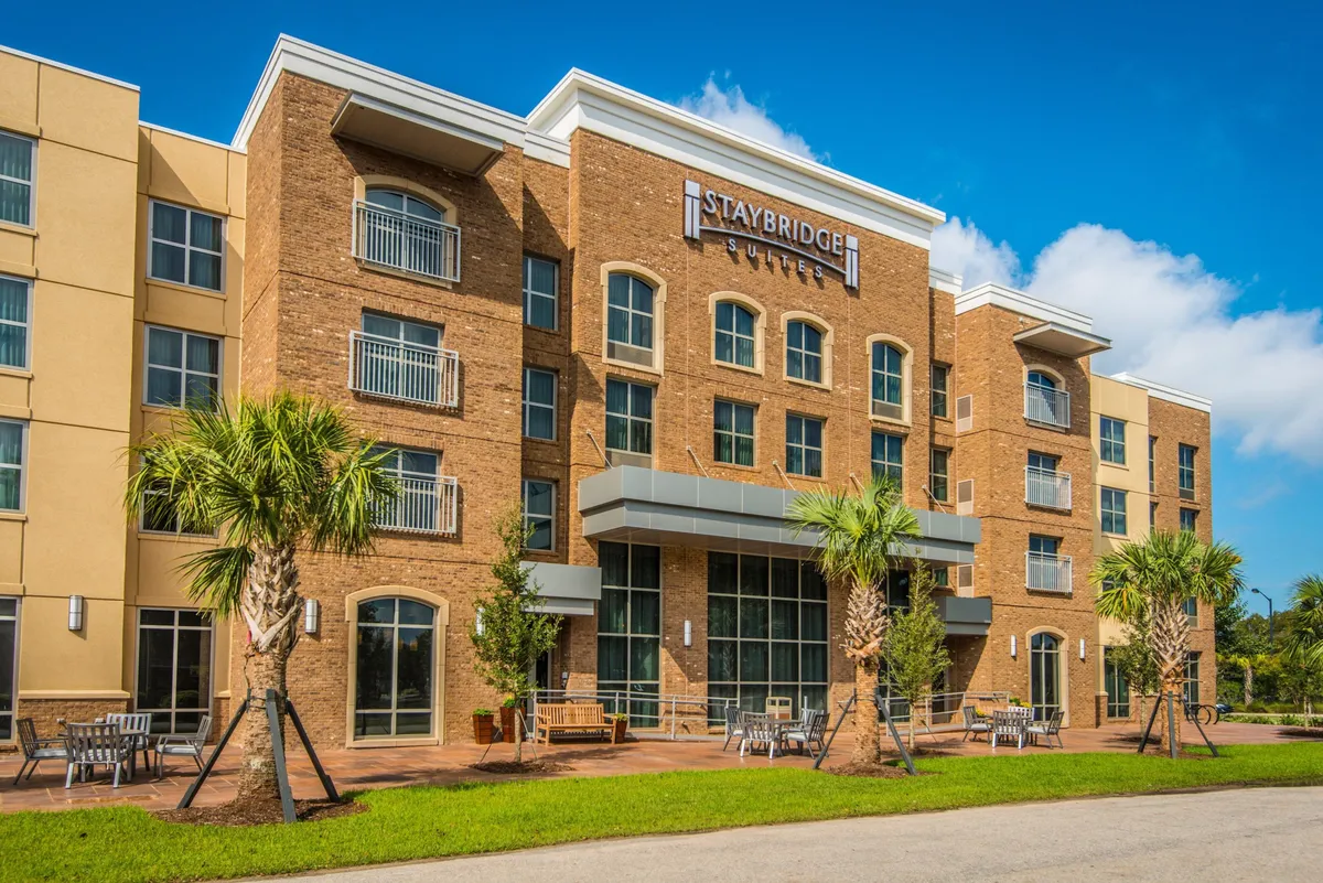 This exterior image features the Staybridge Suites' prominent signage, surrounded by tall palm trees and an inviting outdoor seating area. The scene is bathed in natural light, with guests seen relaxing and enjoying the fresh air, suggesting a connection between indoor productivity and outdoor unwinding.
