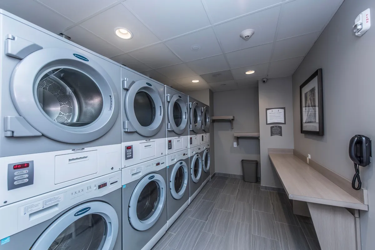 The image presents a practical and tidy laundry room within the hotel, equipped with multiple washing machines, dryers, and ample counter space for folding clothes. The scene conveys ease and convenience for guests caring for their wardrobe during longer stays.
